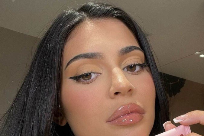 A influencer Kylie Jenner com as sobrancelhas mais finas, os cabelos lisos, compridos e divididos ao meio e com gloss noa lábios