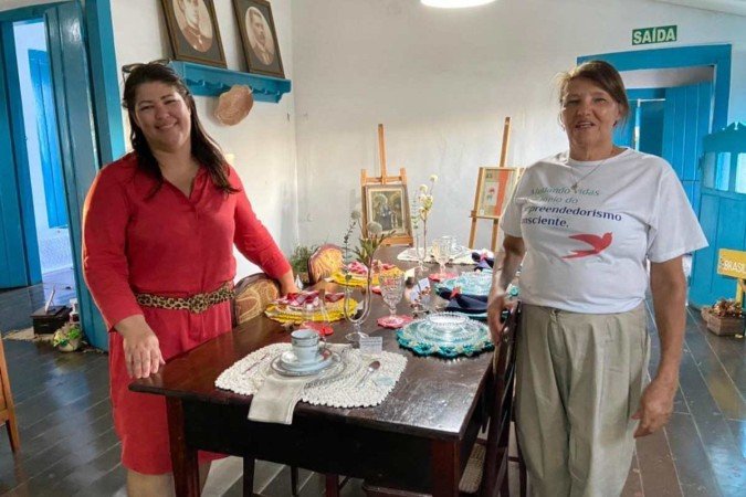 Renata Melo, à esquerda, e Maria Teresinha, à direita, na sala de jantar da exposição Casa de avó. A presidente do Instituto Entre Nós e a coordenadora do projeto comemoram as vendas das peças