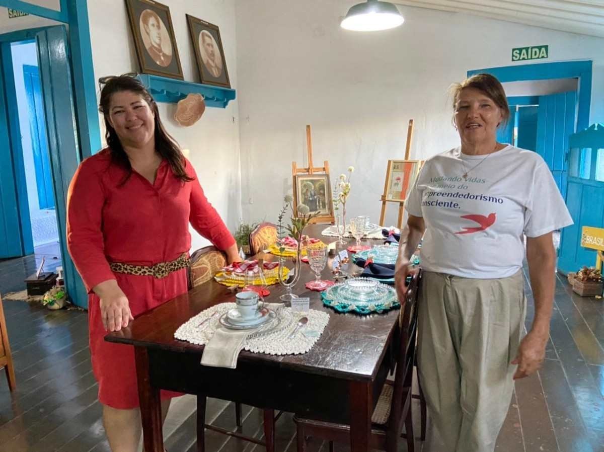 Renata Melo, &agrave; esquerda, e Maria Teresinha, &agrave; direita, na sala de jantar da exposi&ccedil;&atilde;o Casa de av&oacute;. A presidente do Instituto Entre N&oacute;s e a coordenadora do projeto comemoram as vendas das pe&ccedil;as
