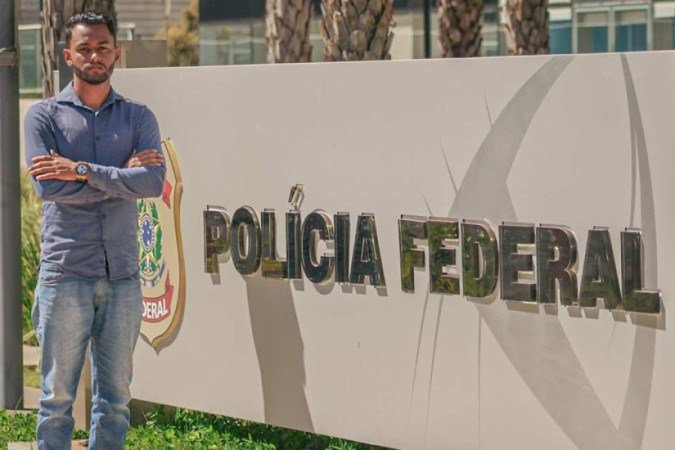 Douglas da Silva Pimenta estuda cerca de 40 horas por semana Homem moreno, de braços cruzados, em pé em frente a um painel da Polícia Federal. Ele veste camisa azul, calça jeans e sapatênis marrom.
