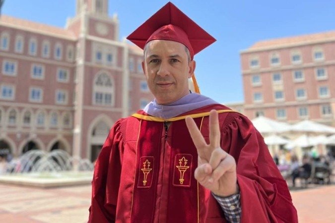 Homem branco com beca vermelha de formando, em frente ao pr&eacute;dio da Universidade do Sul da Calif&oacute;rnia