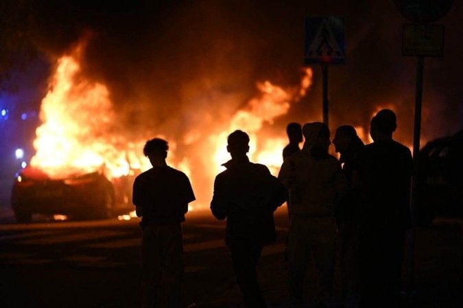Pessoas assistem carros pegando fogo na Ramels vag em Rosengard, Malmo, após manifestação - (crédito: AFP) Pessoas assistem carros pegando fogo na Ramels vag em Rosengard, Malmo, após manifestação - (crédito: AFP)