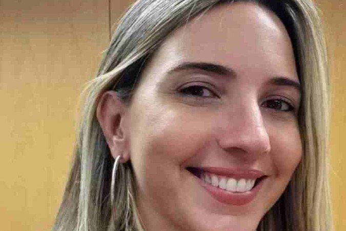 Mulher branca e loira, sorri com uma mão no colo, com um fundo de madeira