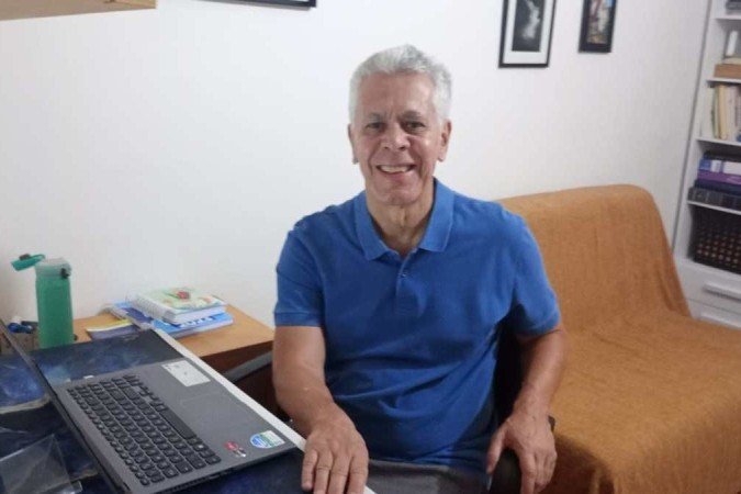 Homem de cabelos grisalhos e camisa azul, posa no escritório de sua casa, em frente ao computador