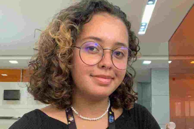 Menina parda de cabelos cacheados posa com o crachá da empresa e escritório ao fundo