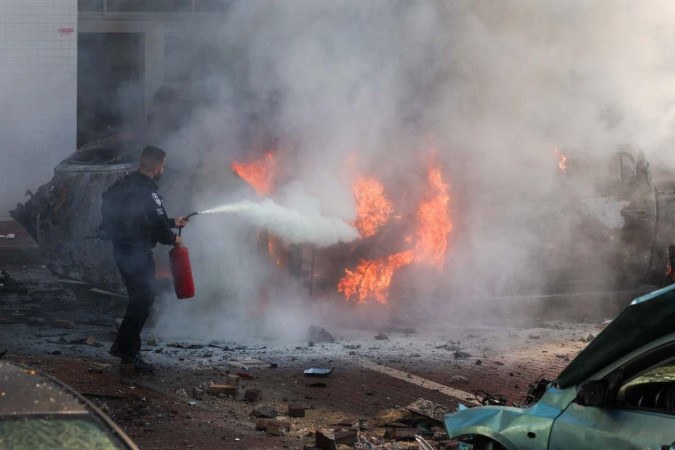       Um membro das forças de segurança israelenses tenta extinguir o fogo contra carros após um ataque com foguetes da Faixa de Gaza em Ashkelon, sul de Israel -  (crédito: Ahmad GHARABLI / AFP)