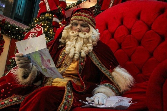 Homem branco fantasiado de papai noel l&ecirc; carta com desenho de crian&ccedil;a sentado em poltrona vermelha.