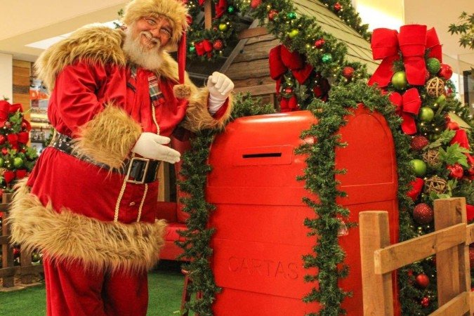 Homem branco fantasiado de papai noel sorri em frente &agrave; casa de madeira cenogr&aacute;fica.