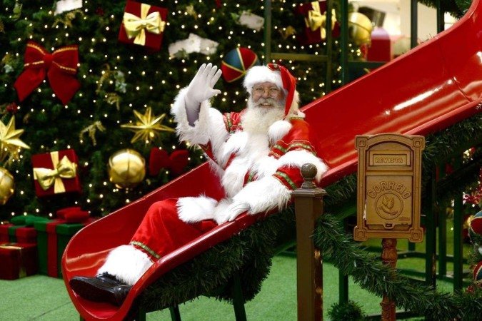 Homem branco fantasiado de papai noel desce escorregador em pra&ccedil;a de shopping com decora&ccedil;&atilde;o de Natal ao fundo.