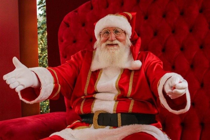 Homem branco fantasiado de papai noel abre os bra&ccedil;os sentado em poltrona vermelha com decora&ccedil;&atilde;o de Natal ao fundo