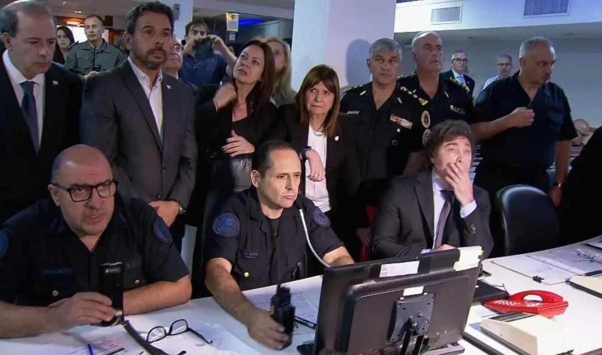 Milei (D, sentado) e a ministra da Seguran&ccedil;a, Patricia Bullrich (C, em p&eacute;), monitoram os protestos em tel&otilde;es da pol&iacute;cia 