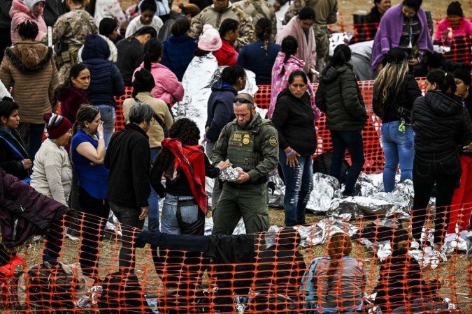 Um oficial da Alfândega e Proteção de Fronteiras dos EUA (C) dá comida a imigrantes que aguardam para serem processados em um centro de trânsito da Patrulha de Fronteira dos EUA, depois de cruzar a fronteira do México em Eagle Pass, Texas, em 22 de dezembro de 2023 - (crédito: CHANDAN KHANNA / AFP) Um oficial da Alfândega e Proteção de Fronteiras dos EUA (C) dá comida a imigrantes que aguardam para serem processados em um centro de trânsito da Patrulha de Fronteira dos EUA, depois de cruzar a fronteira do México em Eagle Pass, Texas, em 22 de dezembro de 2023 - (crédito: CHANDAN KHANNA / AFP)