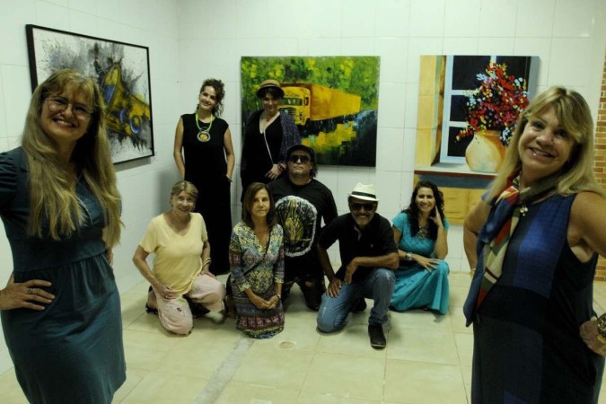 Grupo de nove pessoas posa em galeria de arte, com quadros ao fundo. À frente, estão duas mulheres loiras e brancas.