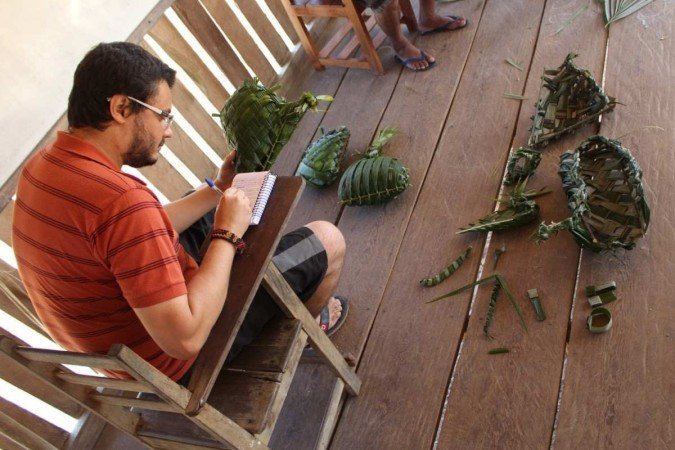 Igor Morais Mariano Rodrigues, professor universitário, foi ganhador do Grande Prêmio Capes na escola de Humanidades Homem branco de óculos sentado em carteira faz anotações em bloco de nota com cestas de folhas ao chão