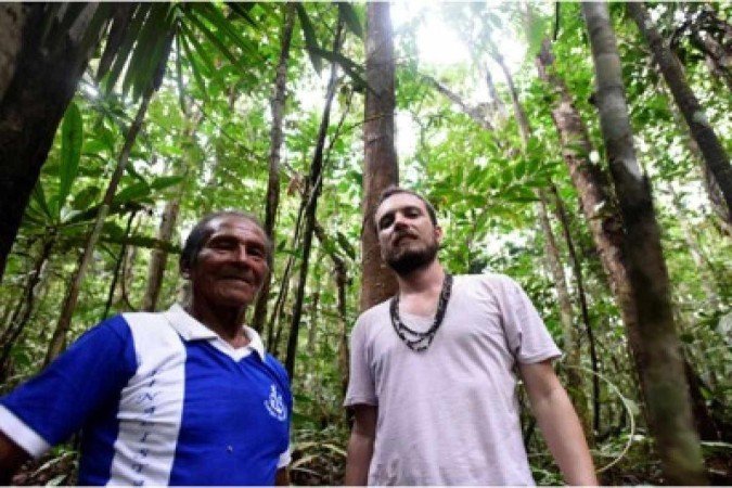 Juliano Franco Morais junto de Pedro Baniwa, líder comunitário do povo Tucumã Rupita Homem branco com camisa branca e colar de sementes posa ao lado de homem indígena de pele escura com camisa polo, floresta ao fundo