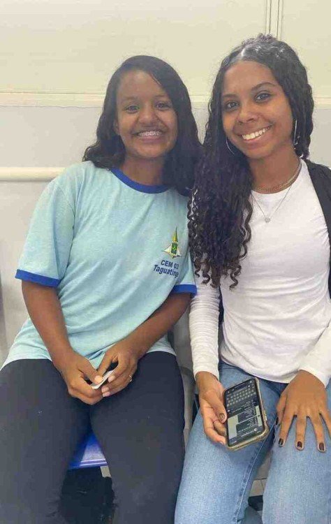 Duas adolescentes negras, de uniforme, sorriem para a foto