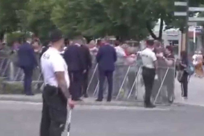 Cercado por seguranças, Robert Fico se aproxima da barrreira; o atirador está de branco (C, ao fundo)