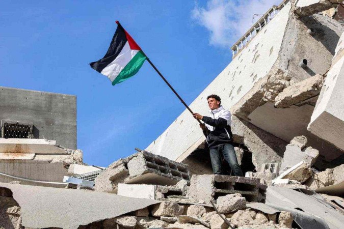     Um homem segura uma bandeira palestina enquanto está no topo dos escombros de um prédio desabado no campo de refugiados palestinos de Jabalia, no norte da Faixa de Gaza, em 19 de janeiro de 2025, após a implementação de um acordo de cessar-fogo na guerra entre Israel e o Hamas. O tão esperado cessar-fogo na guerra Israel-Hamas foi adiado em 19 de janeiro, depois que o primeiro-ministro Benjamin Netanyahu disse no último minuto que não entraria em vigor até que o grupo militante palestino fornecesse uma lista dos reféns a serem libertados. (Foto de Omar AL-QATTAA/AFP)
      