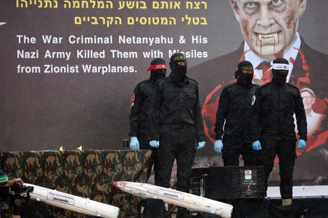Militantes palestinos mascarados ficam ao lado de um dos caixões no palco, com um retrato alterado do primeiro-ministro israelense Benjamin Netanyahu ao fundo, antes de entregar os corpos de quatro reféns israelenses à Cruz Vermelha em Khan Yunis, no sul de Gaza, em 20 de fevereiro - (crédito: Eyad BABA/AFP) Militantes palestinos mascarados ficam ao lado de um dos caixões no palco, com um retrato alterado do primeiro-ministro israelense Benjamin Netanyahu ao fundo, antes de entregar os corpos de quatro reféns israelenses à Cruz Vermelha em Khan Yunis, no sul de Gaza, em 20 de fevereiro - (crédito: Eyad BABA/AFP)