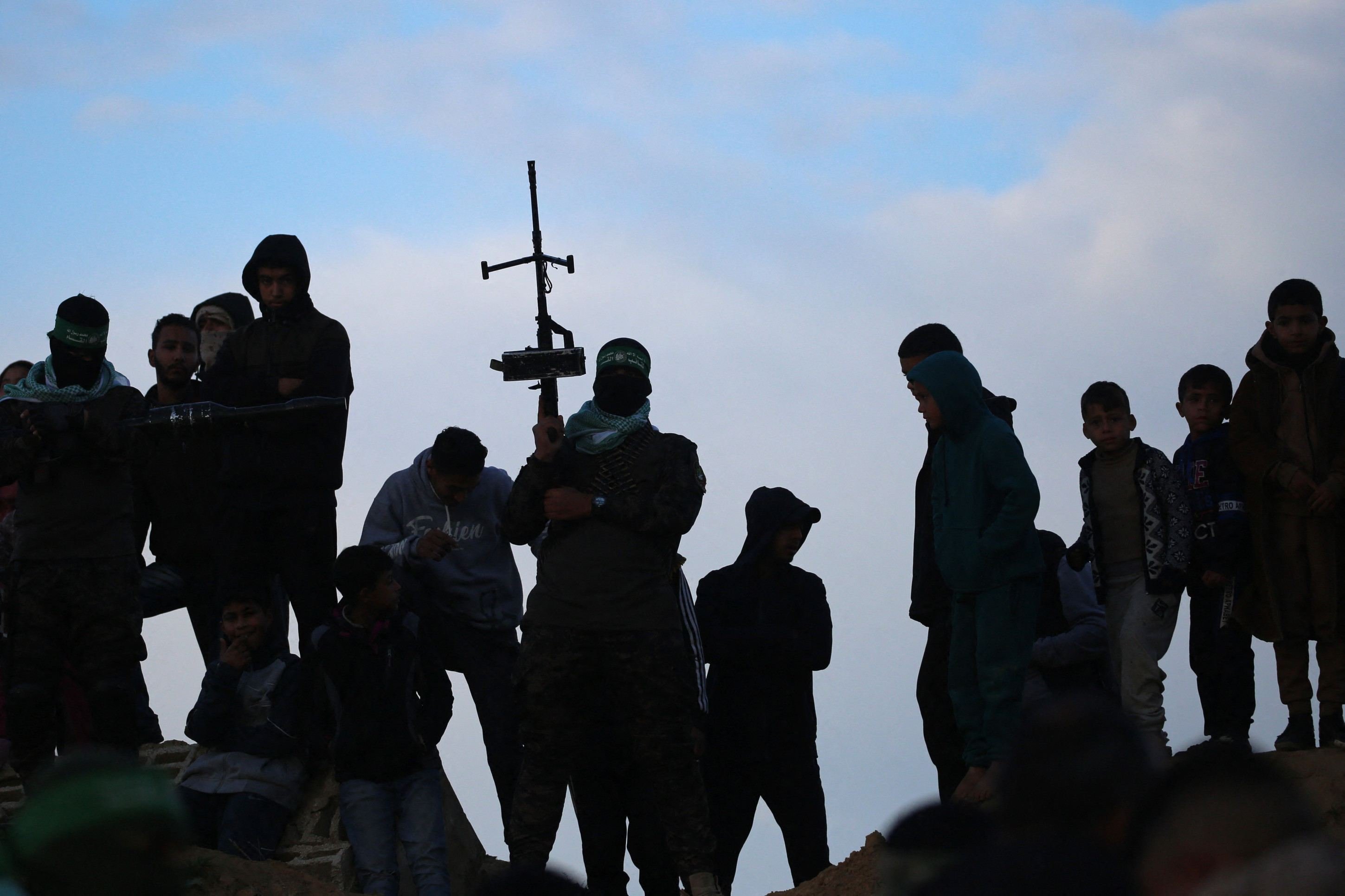 Um militante palestino monta guarda enquanto palestinos estão no telhado de um prédio danificado no local da entrega dos corpos de quatro reféns israelenses em Khan Yunis, no sul de Gaza, em 20 de fevereiro de 2025. O Hamas entregou os corpos de quatro reféns em 20 de fevereiro      