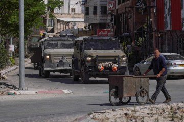 Vendedor ambulante palestino caminha pela rua da mesma cidade, diante de veículos blindados de Israel  