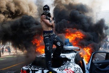 Manifestante mascarado sobre carro autônomo Waymo, depois de atear fogo e pichar o veículo, em Los Angeles, no último domingo  -  (crédito: Blake Flagan/AFP)