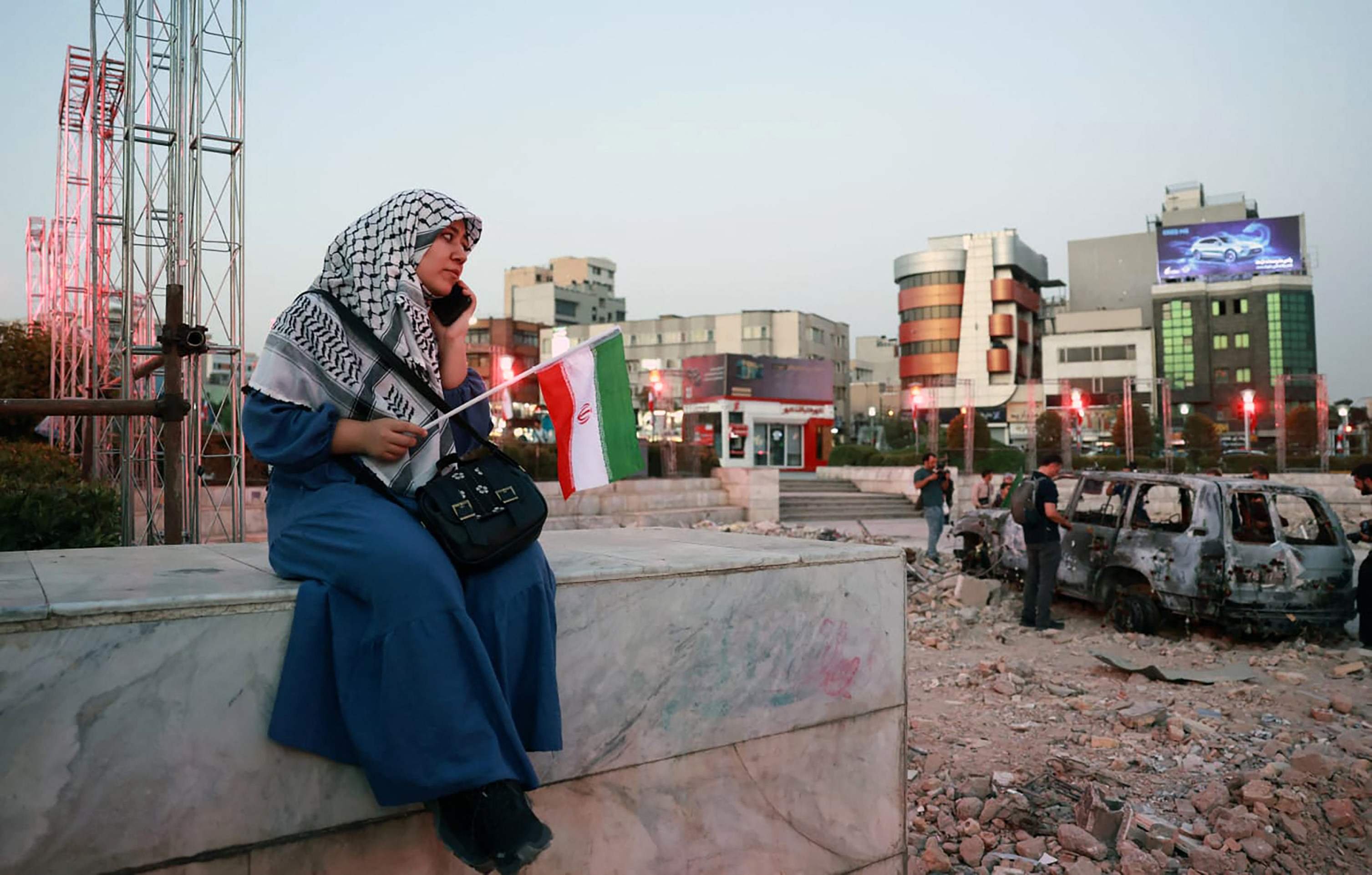 Iraniana senta-se diante de carro destruído, enquanto acompanha exibição de banda tradicional, em homenagem à população, em Teerã