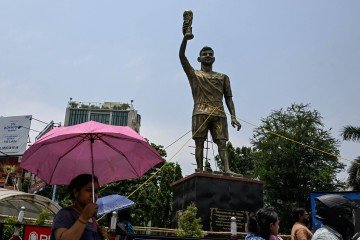 Passageiros passam por uma estátua do jogador de futebol argentino Lionel Messi, que foi amarrada com cordas para maior estabilidade e proteção contra chuva e tempestades, antes de ser reinstalada em um local mais seguro, em Calcutá, em 27 de maio de 2026       -  (crédito: DIBYANGSHU SARKAR / AFP)