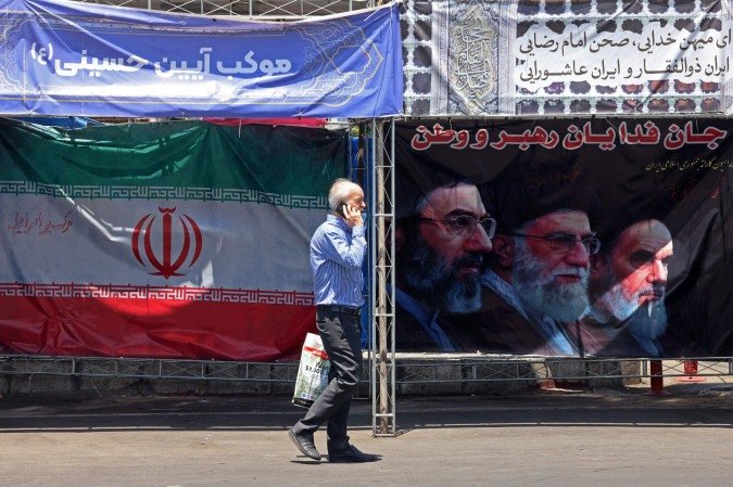 Um homem caminha ao lado da bandeira nacional do Irã e de uma faixa com as imagens do falecido fundador da Revolução Islâmica, o líder supremo Aiatolá Ruhollah Khomeini (à direita), do falecido líder supremo iraniano Ali Khamenei e de seu filho, o atual líder supremo Mojtaba Khamenei (à esquerda), erguidas ao longo de uma rua na Praça Vanak, em Teerã, em 10 de junho de 2026 - (crédito: ATTA KENARE / AFP) Um homem caminha ao lado da bandeira nacional do Irã e de uma faixa com as imagens do falecido fundador da Revolução Islâmica, o líder supremo Aiatolá Ruhollah Khomeini (à direita), do falecido líder supremo iraniano Ali Khamenei e de seu filho, o atual líder supremo Mojtaba Khamenei (à esquerda), erguidas ao longo de uma rua na Praça Vanak, em Teerã, em 10 de junho de 2026 - (crédito: ATTA KENARE / AFP)