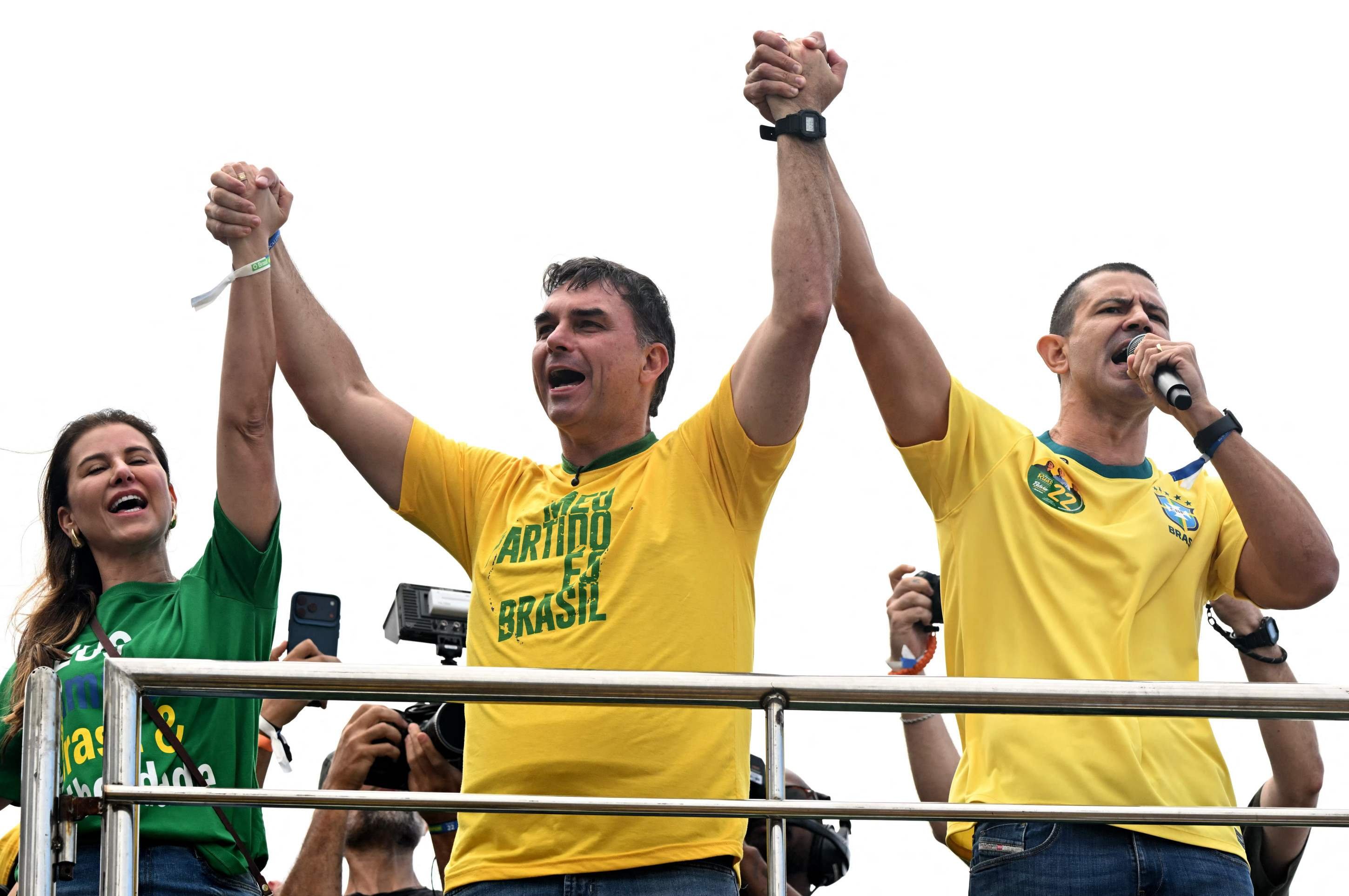  Brazil's right-wing presidential candidate and senator Flavio Bolsonaro (C) holds hands with his wife Fernanda Bolsonaro and his candidate for Rio de Janeiro governor Douglas Ruas during a campaign rally at Copacaban beach in Rio de Janeiro, Brazil, on August 16, 2026. Brazilians will go to the polls next October 4 once again deeply polarized and under the shadow of U.S. President Donald Trump, who has backed conservative candidates in recent elections in Latin America. (Photo by MAURO PIMENTEL / AFP)       